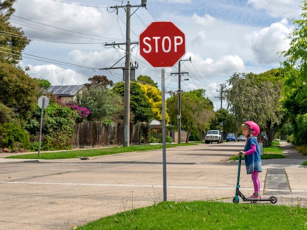 A little girl crosses the street on an e-scooter at a stop sign